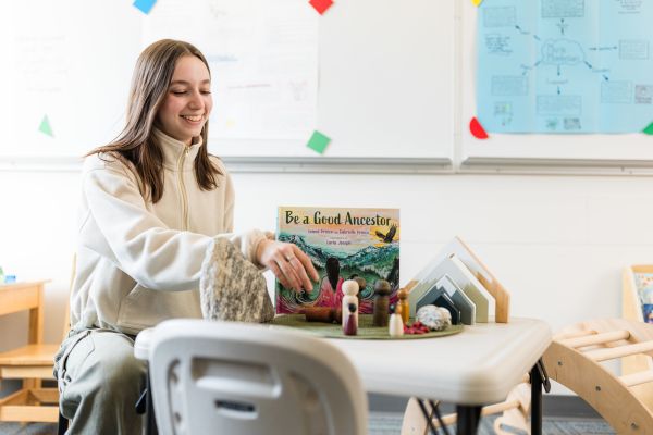 Female student with brown hair sits at a table with a children's book and some toys.