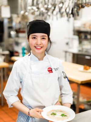 A female Culinary Arts student smiles as she holds a garnished bowl of soup.