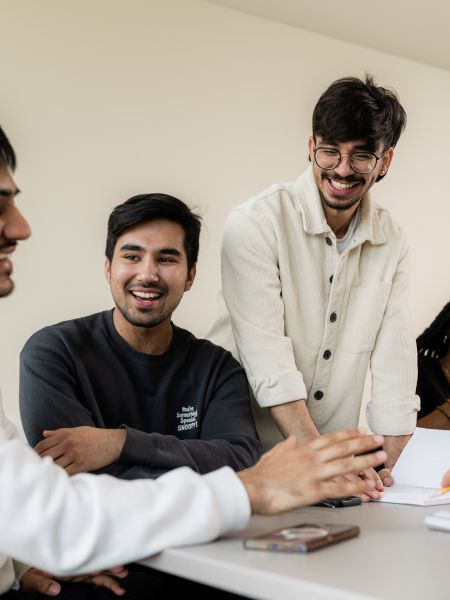 A group of five college students sit and stand around a table, smiling and laughing together during a casual study or discussion session. Notebooks and a phone are on the table, and the mood is friendly and collaborative.