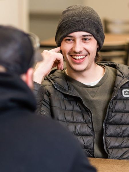 Student in a hat and jacket sitting at a table, smiling while facing another student sitting in the foreground with their back to the camera.