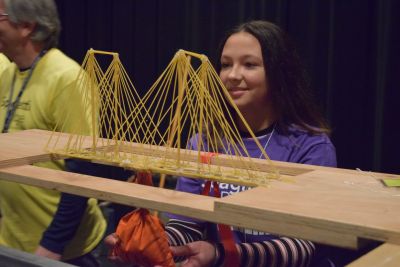 a student behind a cable-stayed style bridge made out of spaghetti at the Spaghetti Bridge Building Contest
