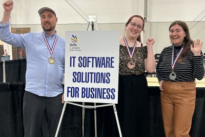 Three individuals stand on a podium indoors, each wearing medals and raising an arm in celebration. A sign reads "IT SOFTWARE SOLUTIONS FOR BUSINESS."
