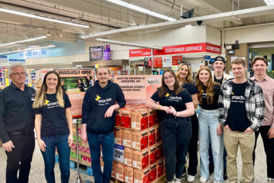 Eight people, some in "enactus" shirts, stand in a grocery store beside a product display promoting donations of meals to Canadian schools and food banks.