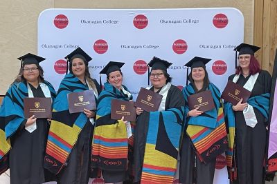 Group of graduates in caps and gowns holding diplomas and wearing colorful ceremonial blankets, standing in front of an Okanagan College backdrop during a graduation event