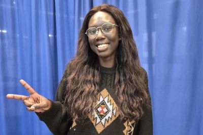 Person standing in front of a blue curtain wearing a dark knitted sweater, holding up a peace sign with the left hand.