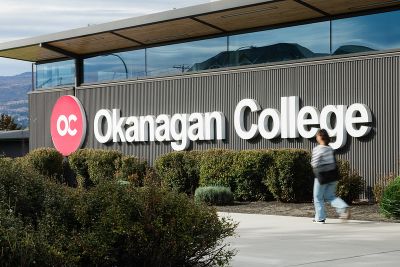 building with the text "Okanagan College", a woman walking on the sidewalk in front of building