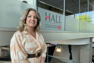 Woman standing indoors near a glass railing in front of a “Hall School of Business and Entrepreneurship” sign, wearing a cream floral blouse, brown belt, and large gold statement earrings, smiling at the camera.