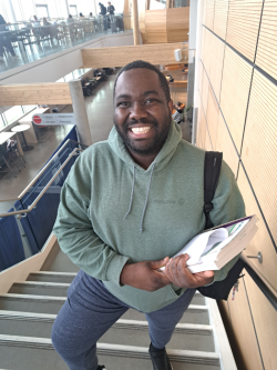smiling student holding books and a backpack, wearing a green sweatshirt and blue pants.