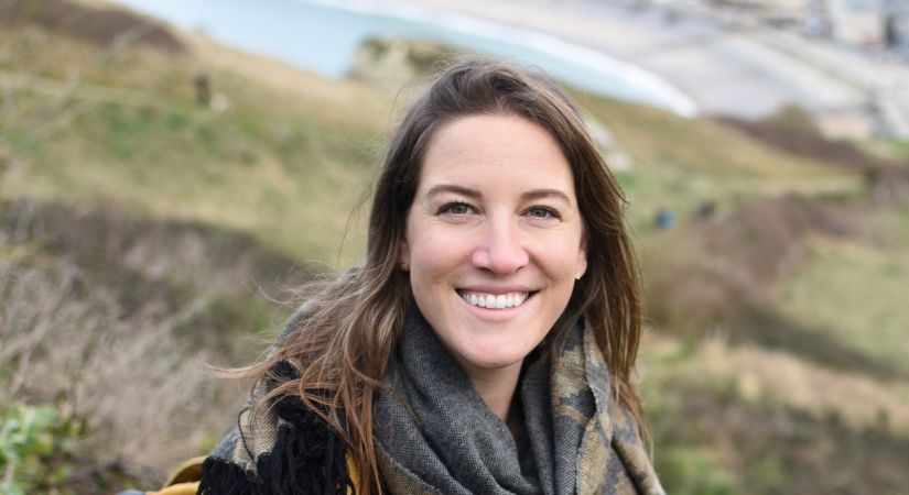 A female stands on a hill outside in a jacket and scarf wearing a backpack.