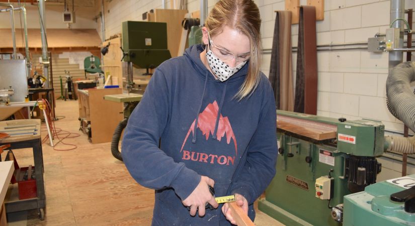 Female carpentry student measures a piece of wood with a tape measure
