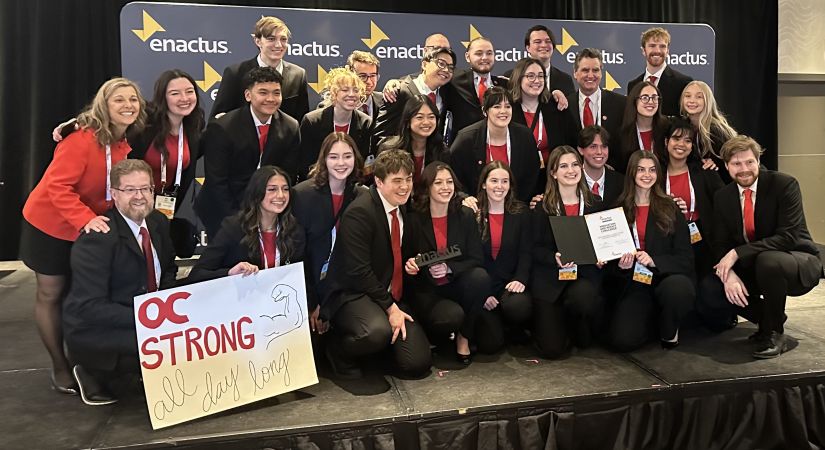 Enactus group photo. Students smiling together while on stage holding a sign that reads "O C strong, all day long"