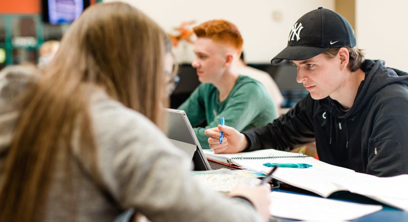 Three students are sitting at a table, working on their laptops and notebooks in a study environment.