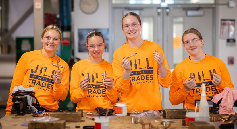 Four individuals in a workshop, wearing bright orange shirts with "JILL OF ALL TRADES A DAY FOR WOMEN" printed on them, hold tools while standing in front of a table with various materials.