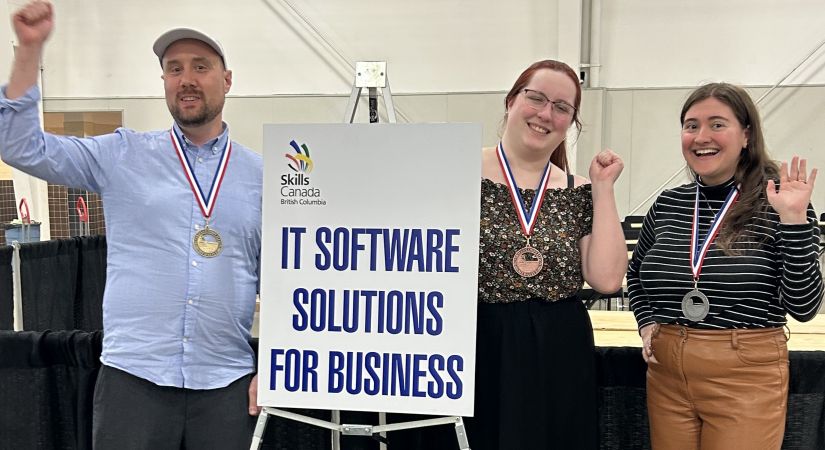 Three individuals stand on a podium indoors, each wearing medals and raising an arm in celebration. A sign reads "IT SOFTWARE SOLUTIONS FOR BUSINESS."