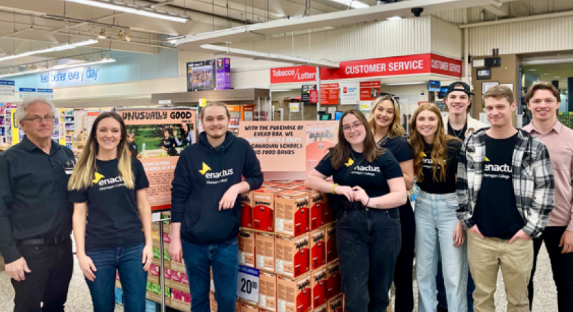 Eight people, some in "enactus" shirts, stand in a grocery store beside a product display promoting donations of meals to Canadian schools and food banks.