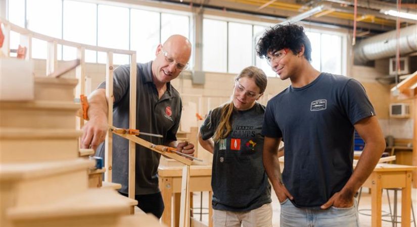 Three people in a woodworking workshop, with one demonstrating a measuring tool near a wooden staircase while the others observe.