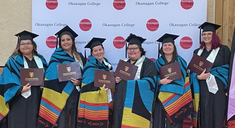 Group of graduates in caps and gowns holding diplomas and wearing colorful ceremonial blankets, standing in front of an Okanagan College backdrop during a graduation event