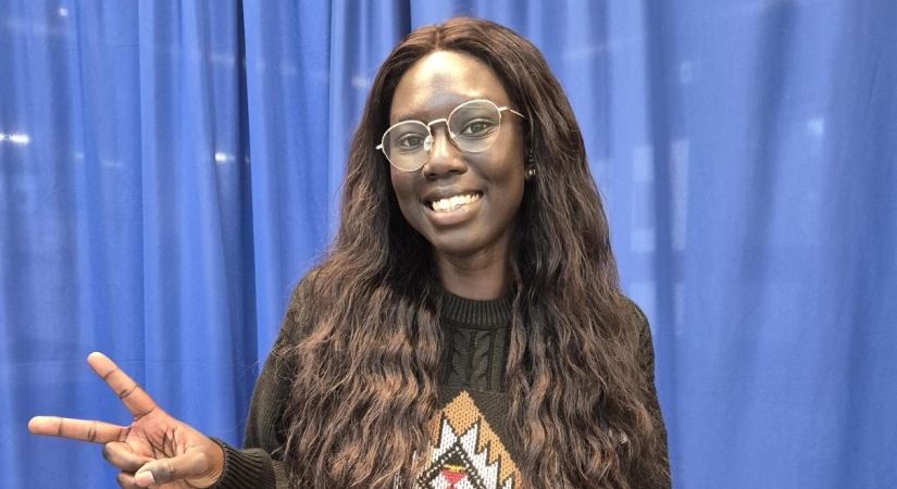 Person standing in front of a blue curtain wearing a dark knitted sweater, holding up a peace sign with the left hand.