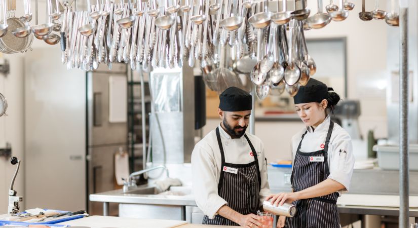 two culinary students stand in a kitchen pouring a drink
