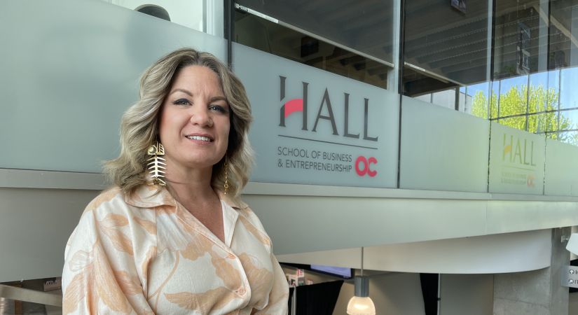 Woman standing indoors near a glass railing in front of a “Hall School of Business and Entrepreneurship” sign, wearing a cream floral blouse, brown belt, and large gold statement earrings, smiling at the camera.